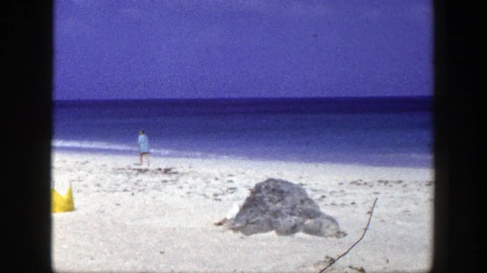 8mm beach frame with the man walking left of the rock, a partial yellow tent, pale sand, blue sea, and visible film texture.