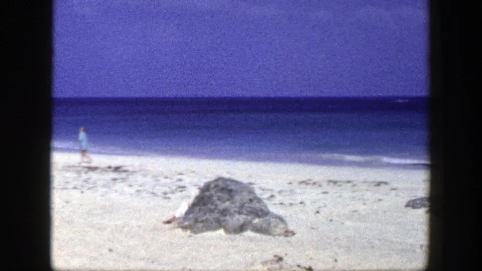 1948 home-movie shoreline frame with a lone man near the surf, a large beach rock, white sand, blue water, and saturated sky.