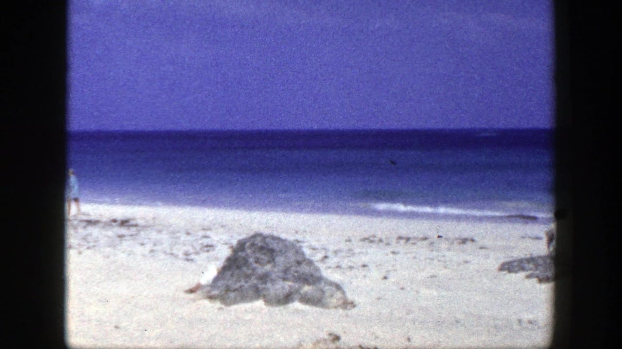Pale sandy beach with rocks, calm ocean, and a small seated figure near the shore.