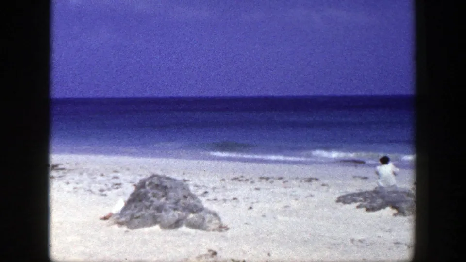 1948 New York beach frame with a seated person on a low rock, calm waves, pale sand, and scattered shoreline rocks.