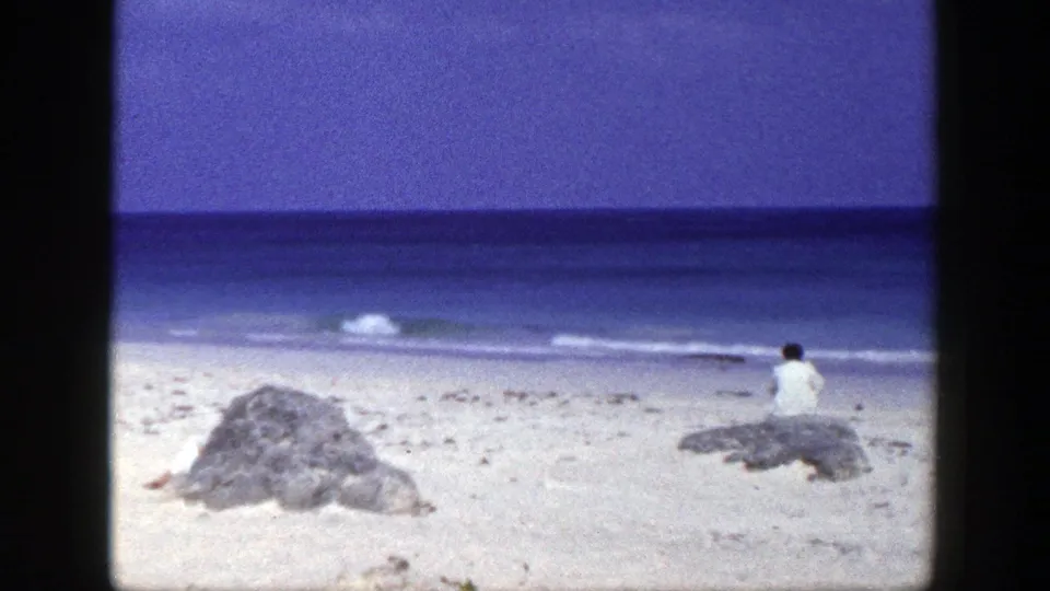 1948 New York 8mm still showing a quiet beach, a foreground rock, calm ocean, and a person seated near the water.