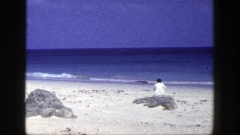 Late-1940s New York beach frame with a person seated on a shoreline rock, calm surf, pale sand, and saturated blue water.