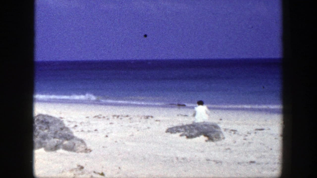 Woman in white seated on a beach rock facing the ocean under a blue sky.