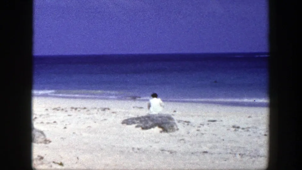 1948 New York beach frame showing a woman in white seated near shoreline rocks with the ocean in the background.