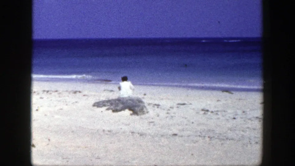 1948 New York 8mm frame of a solitary woman on a sandy beach, seated by rocks and facing the ocean.