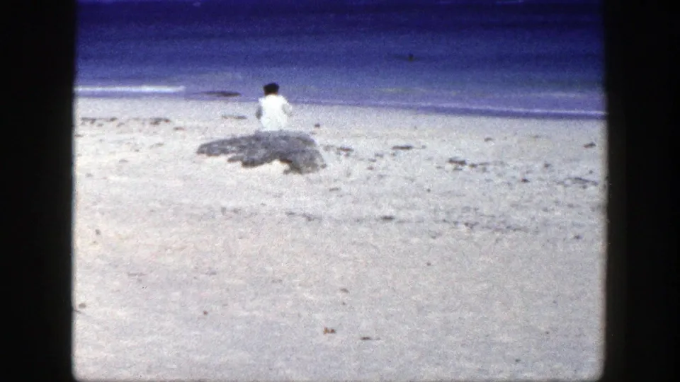 1948 New York beach frame with a woman in white seated beside rocks, looking out toward the blue ocean.
