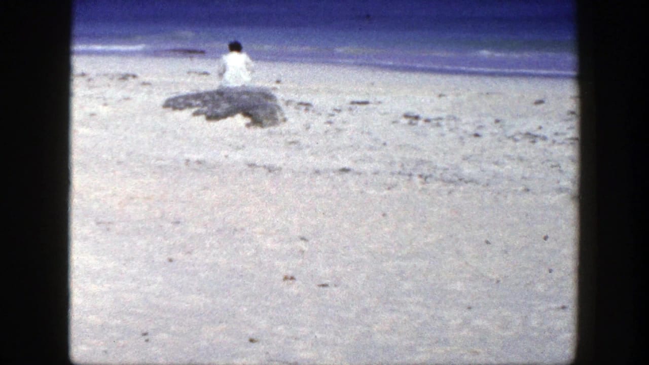 Person in a white shirt sits on a rock near a sandy beach and blue water.