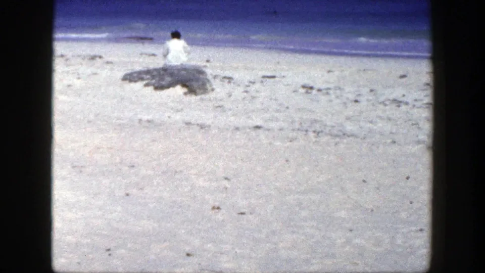 1948 New York home-movie frame showing a distant person seated on a shoreline rock beside sand and calm blue water.
