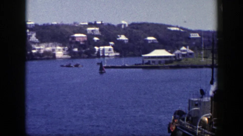 Mid-century New York home-movie frame with boats on calm water, a shoreline structure, and hillside houses in the distance.