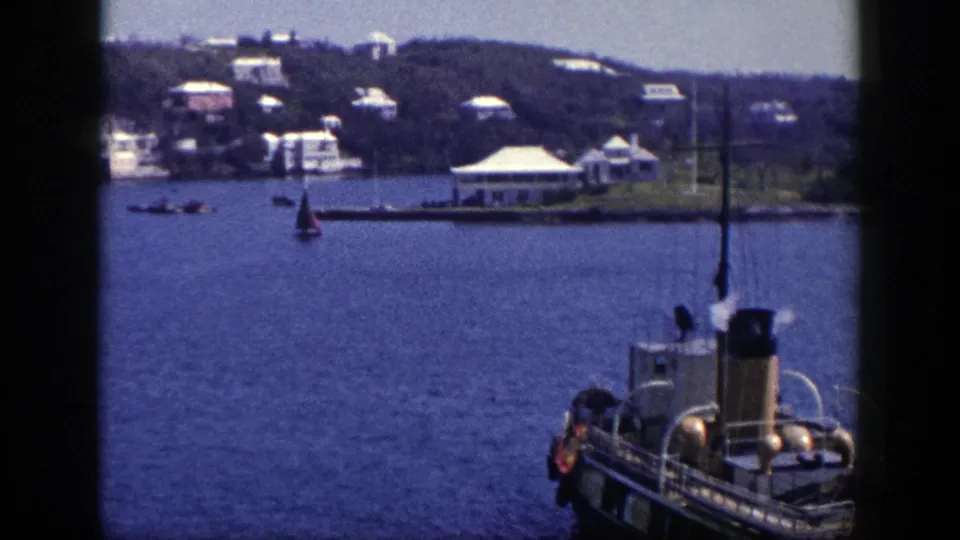Home-movie frame of a New York harbor scene, showing a steam boat foreground, a small sailboat, and white buildings along the far shore.