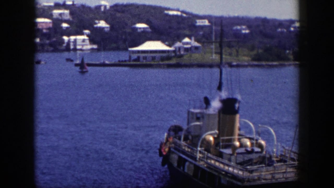 Steam boat on blue harbor water with shoreline buildings and a small sailboat behind it.