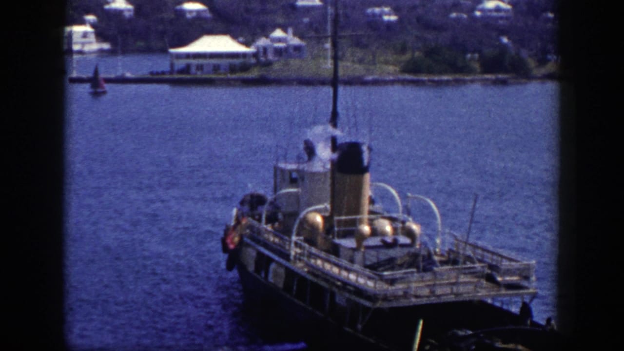 Dark-hulled steamboat on blue water with shoreline buildings behind it.