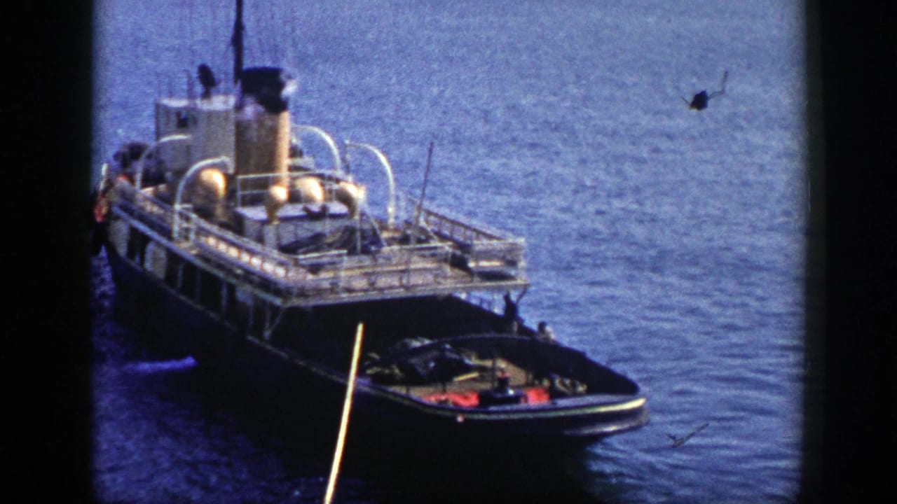 Tugboat seen from above on calm blue water with a smoke plume and small bow boat.