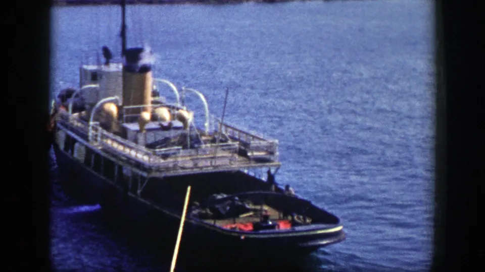 1948 New York 8mm frame of a tugboat with white rails, a yellow stack releasing smoke, and a small boat positioned near the bow.