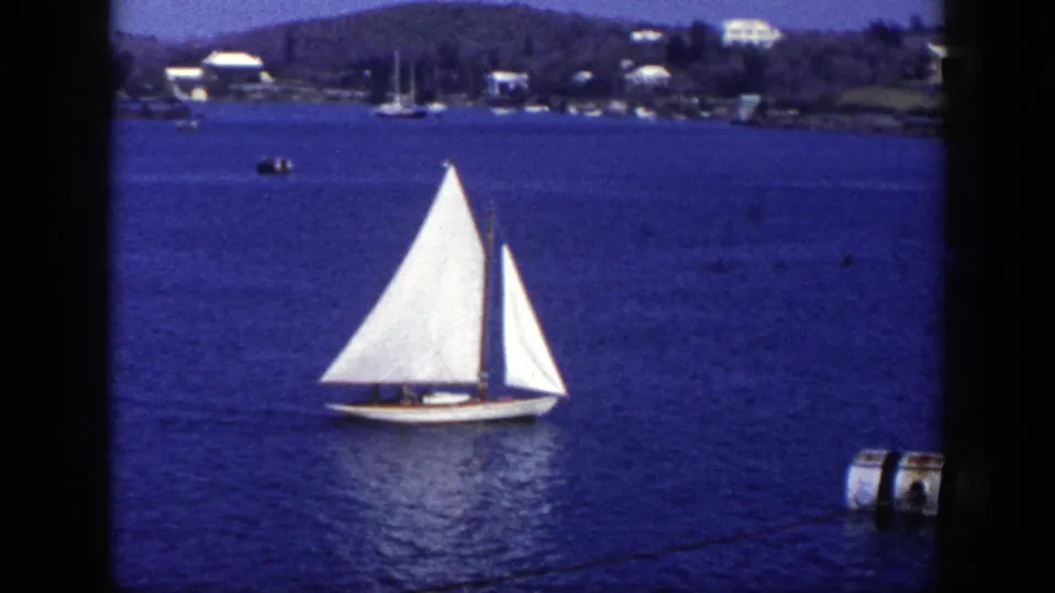 1948 New York 8mm frame of a white sailboat on blue harbor water, with houses and trees along the distant shore.
