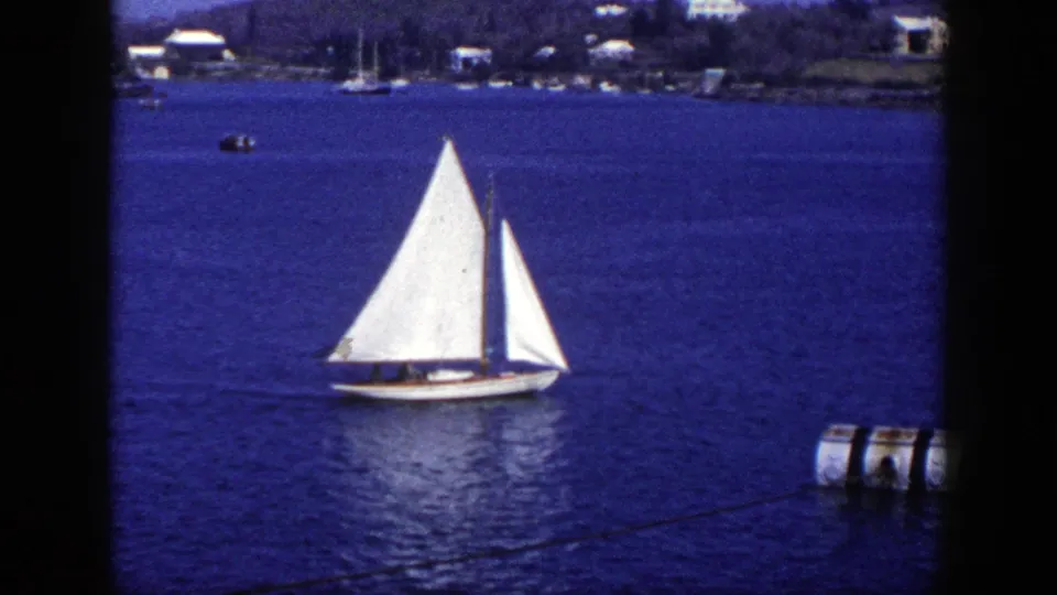 1948 New York 8mm frame of a white sailboat on harbor water, with distant shore houses and dock floats at right.