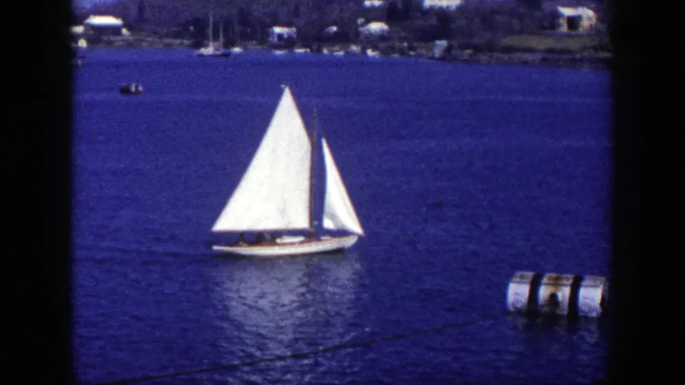 1948 New York 8mm frame of a white sailboat gliding across blue harbor water, with small figures aboard and shoreline details in the background.