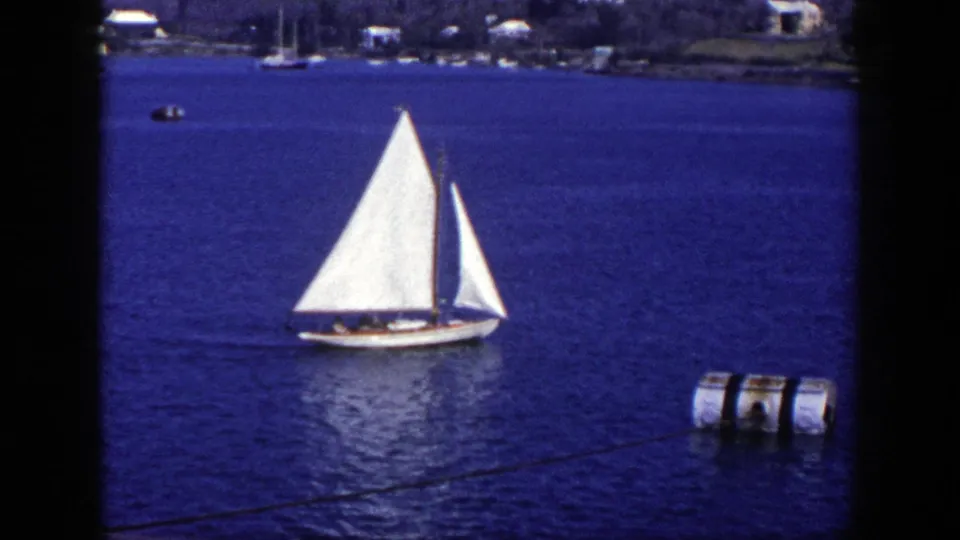 1948 New York home-movie frame showing a white sailboat on blue water, distant moored boats, shoreline houses, and a barrel-like floating object at right.