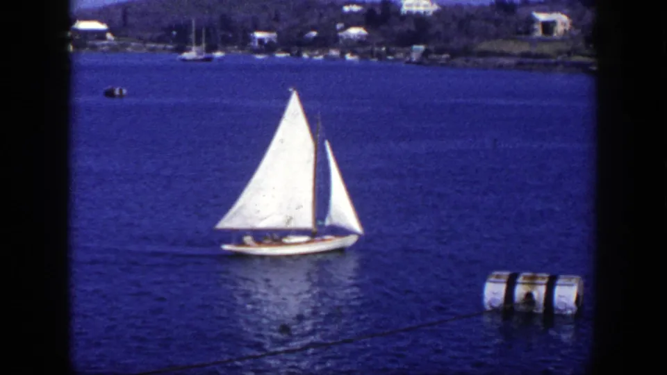 1948 New York 8mm frame of a white sailboat moving across deep blue water, with tiny people aboard, distant shore, and floating object at the edge.