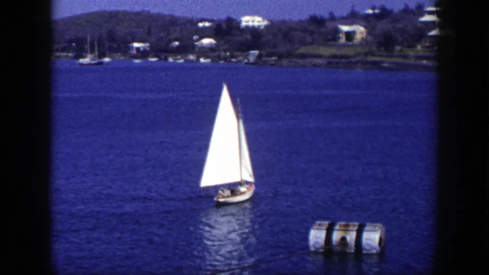 1948 New York waterfront frame showing a small sailboat turning or passing near a cylindrical buoy, with shoreline homes in the distance.