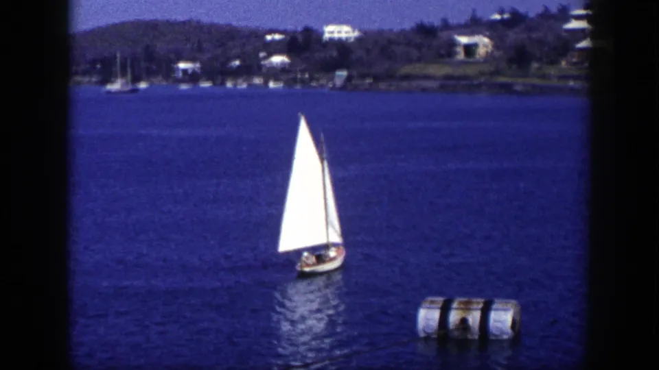 1948 New York 8mm frame showing a small sailboat on bright blue water, a floating cylindrical buoy, and hillside houses in the distance.