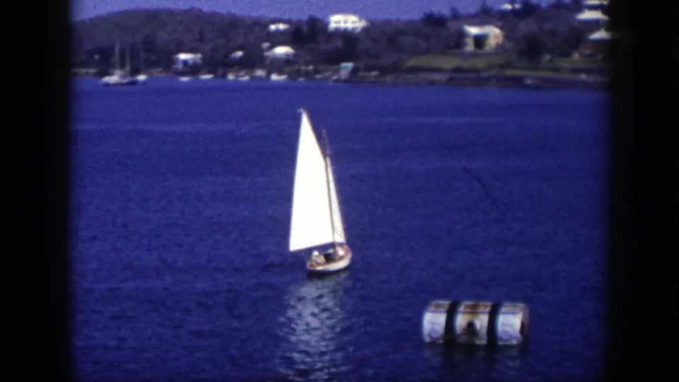 1948 New York 8mm frame showing a white-sailed boat moving across blue water with a buoy and residential wooded shoreline beyond.