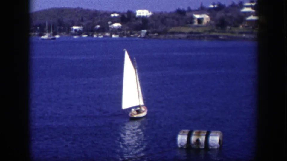 1948 New York home-movie frame of a small sailboat on bright water, with a cylindrical buoy, moored boats, trees, and hillside homes behind.