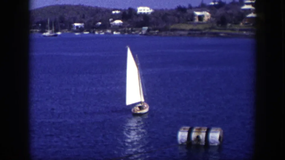1948 New York 8mm frame of a white-sailed boat moving across blue water near a buoy, with trees, houses, and moored boats along the far shore.