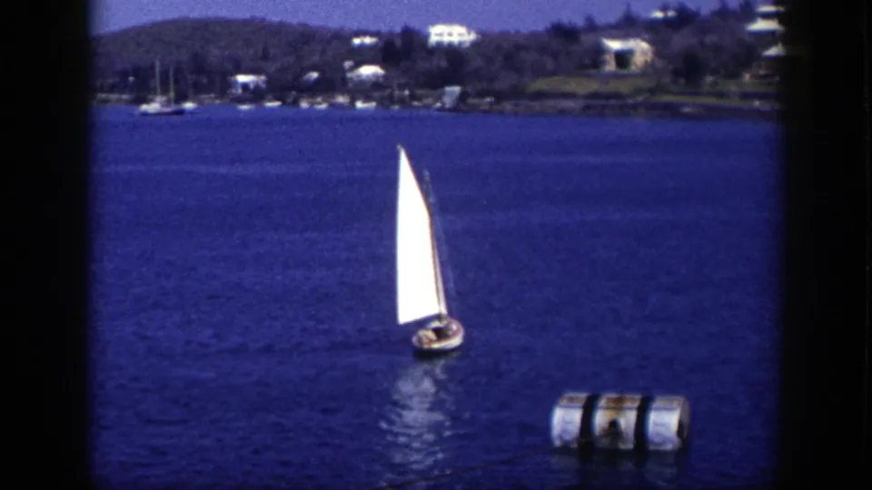 1948 New York 8mm frame showing a small sailboat with a bright white sail, a nearby cylindrical buoy, and a residential shoreline in the background.
