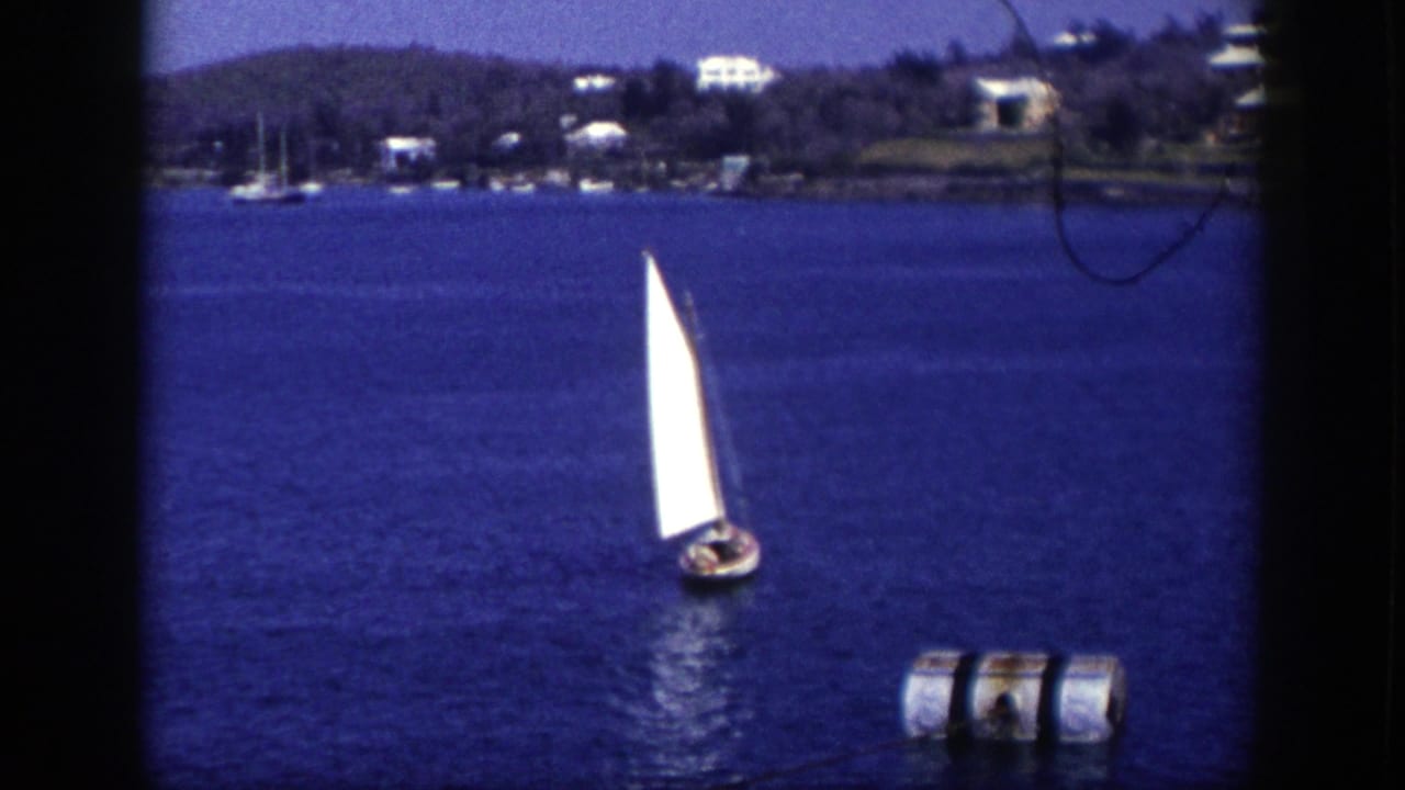 1948 New York 8mm frame of a small sailboat crossing deep blue coastal water, with a buoy in front and houses along the far shore.