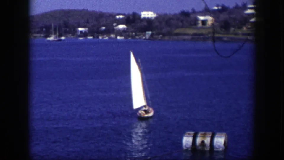 1948 New York 8mm frame of a small sailboat crossing deep blue coastal water, with a buoy in front and houses along the far shore.