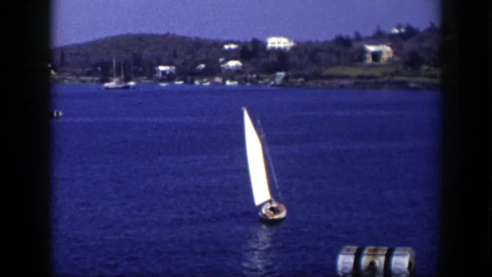 1948 New York 8mm frame of a white-sailed boat and its reflection on coastal water, with a buoy and distant hillside homes visible.