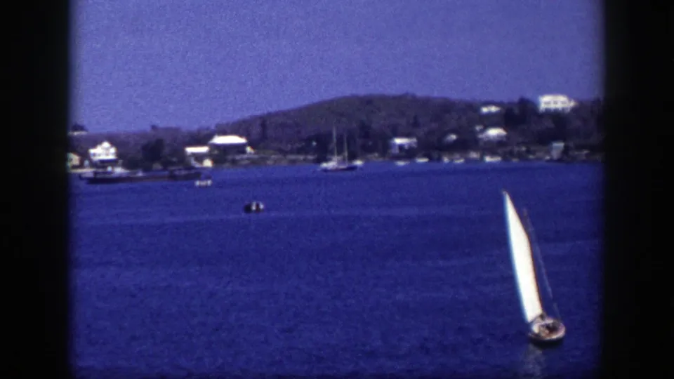 1948 New York home-movie frame with a white sailboat on open blue water, backed by small boats, houses, trees, and low hills.