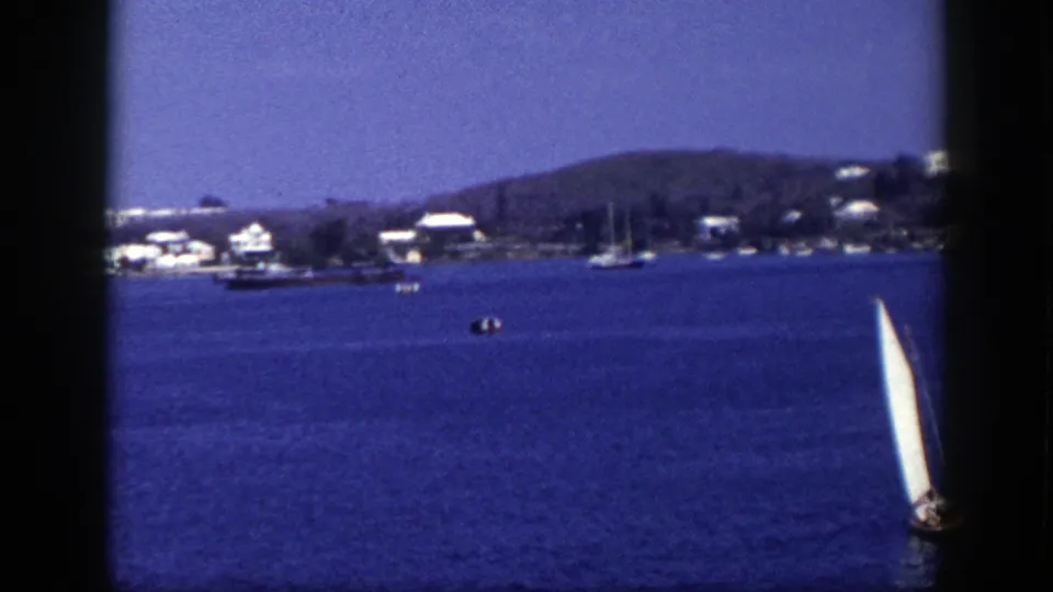 1948 New York 8mm frame looking across blue water toward shoreline houses, moored boats, and a partly visible white sailboat.