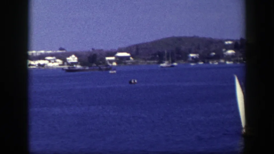 1948 New York home-movie frame with open blue water, distant shoreline homes, moored boats, and the sailboat nearly leaving the frame.