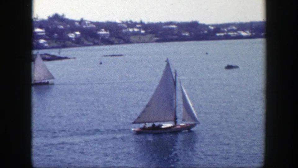 A white-sailed boat crosses calm New York harbor water in this 1948 home-movie frame, with shoreline houses behind it.