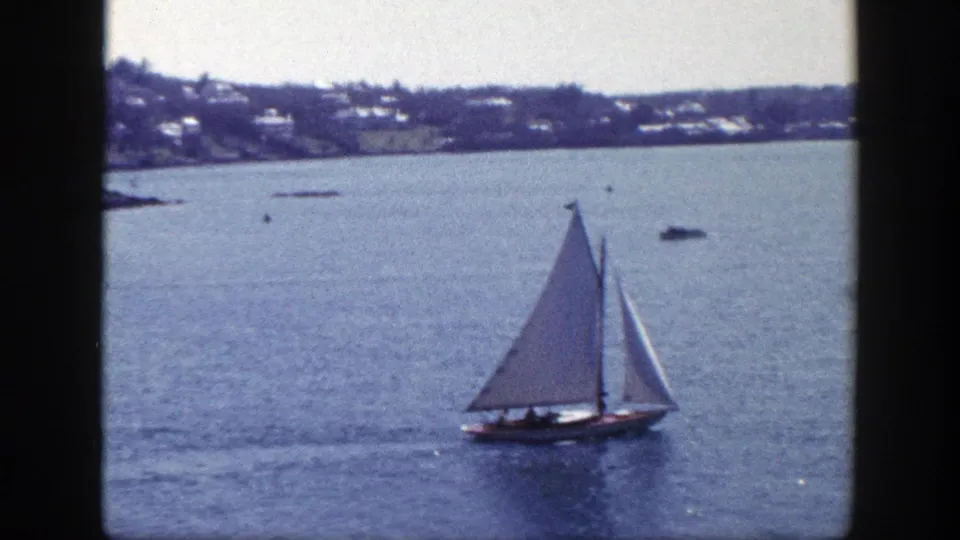 White sails and a dark hull stand out on calm New York water in this 1948 home-movie frame, with the shoreline beyond.