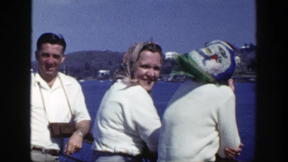 In 1948 New York 8mm footage, three adults in white clothing stand near a waterfront railing with distant shoreline behind them.