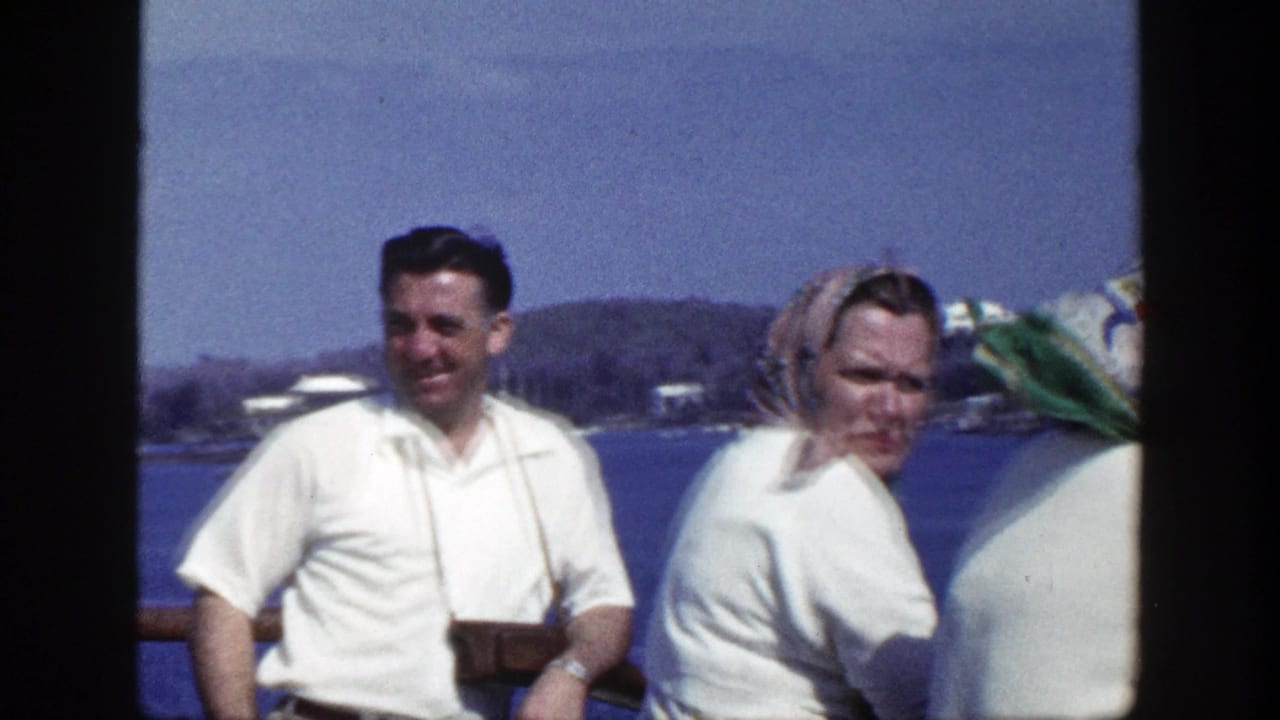 A smiling man with a camera stands beside two women in white sweaters near blue water.