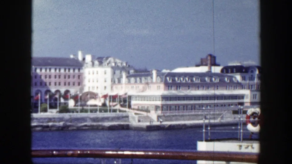 1948 New York waterfront buildings with arched openings and red flags viewed across harbor water from a ship railing.