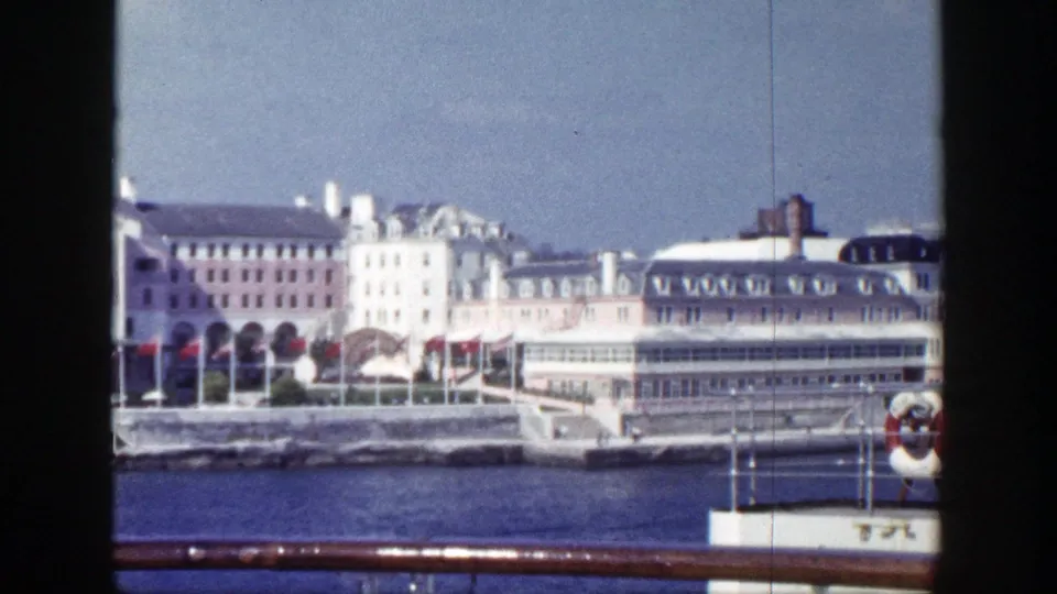 A shipboard 8mm view of New York waterfront buildings, red flags, and calm harbor water under blue sky.
