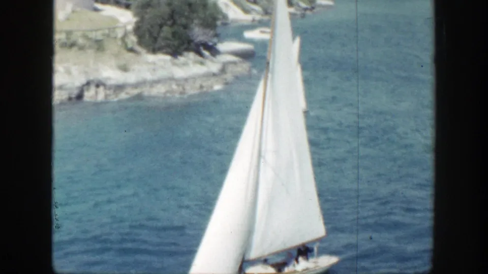 1948 New York 8mm frame showing a white sailboat on calm blue water near rocks and trees along the shore.