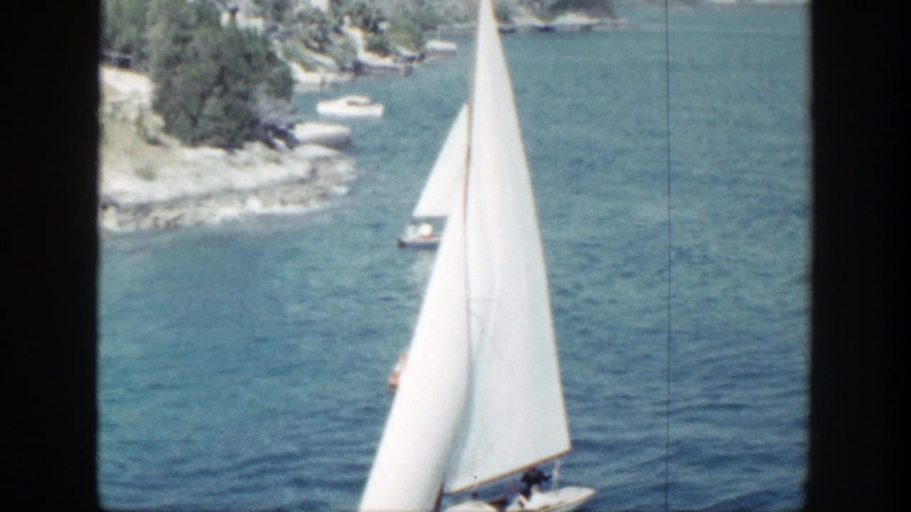 White sailboat on blue water with shoreline and small boats behind it.