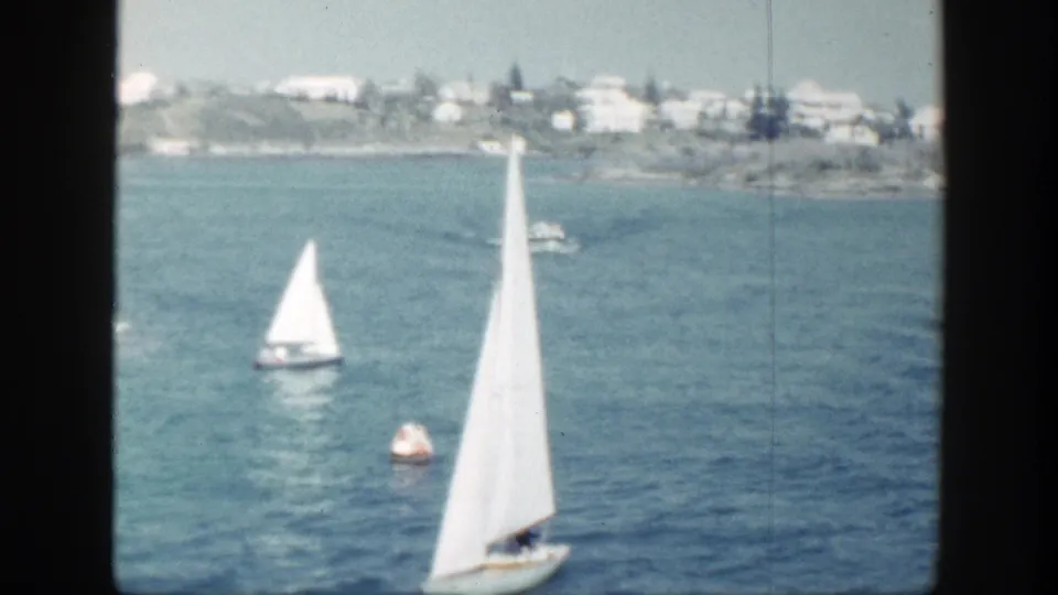 Two white sailboats and a buoy appear on blue coastal water, with a distant town shoreline in the background.