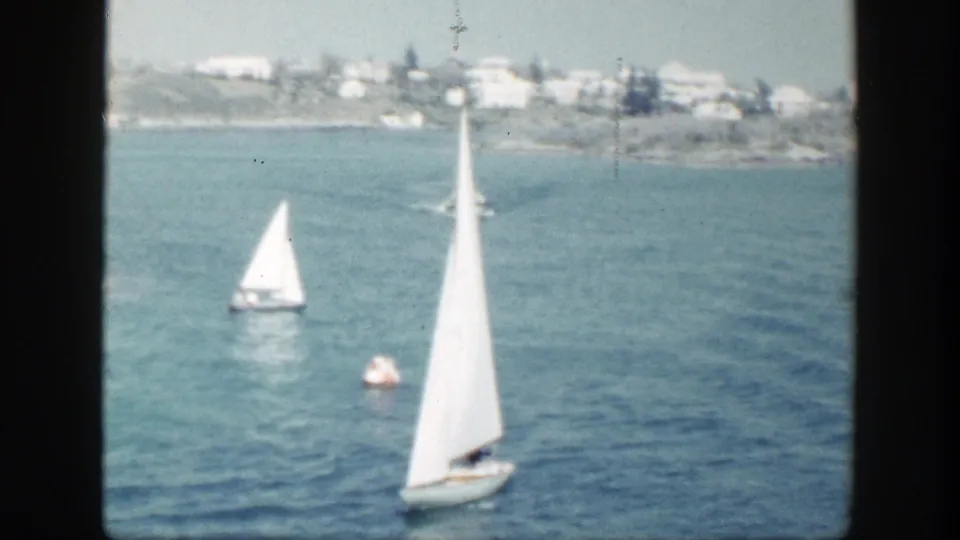 Multiple sailboats move across calm New York coastal water, with a buoy and distant shoreline buildings beyond.