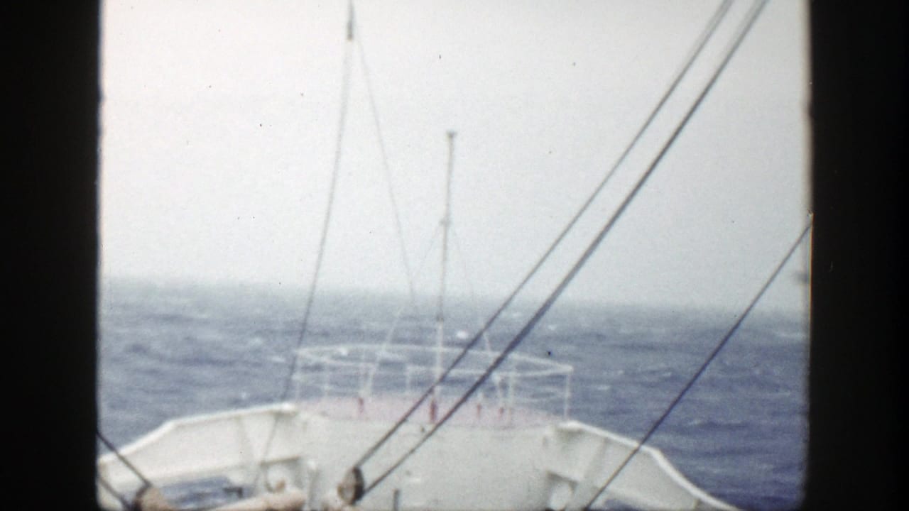 1948 home-movie frame of a vessel bow, railings, and rigging facing rough open water under a pale sky.
