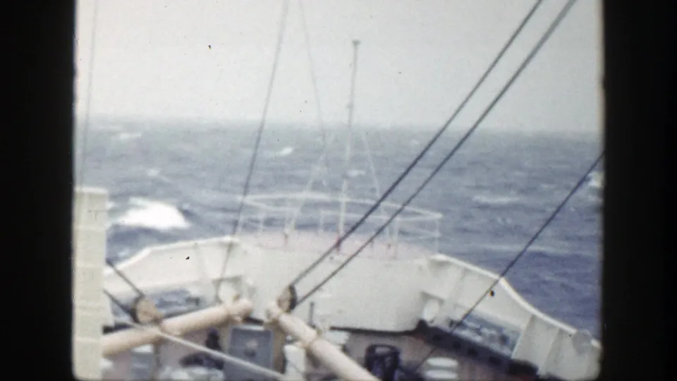 A ship’s forward deck and rigging are seen as white water breaks along the hull in vintage 8mm footage.