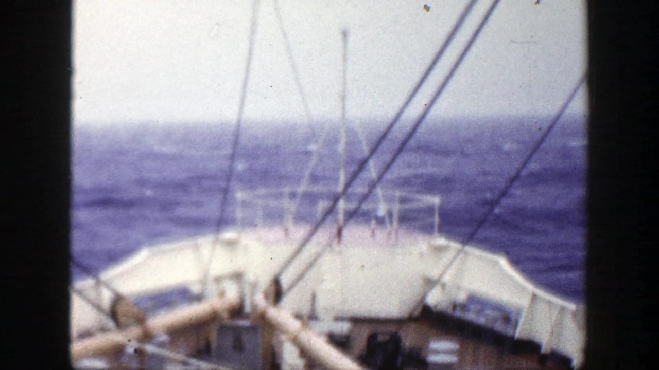 1948 8mm deck view showing the ship bow, railings, and rigging against rough blue sea and pale sky.
