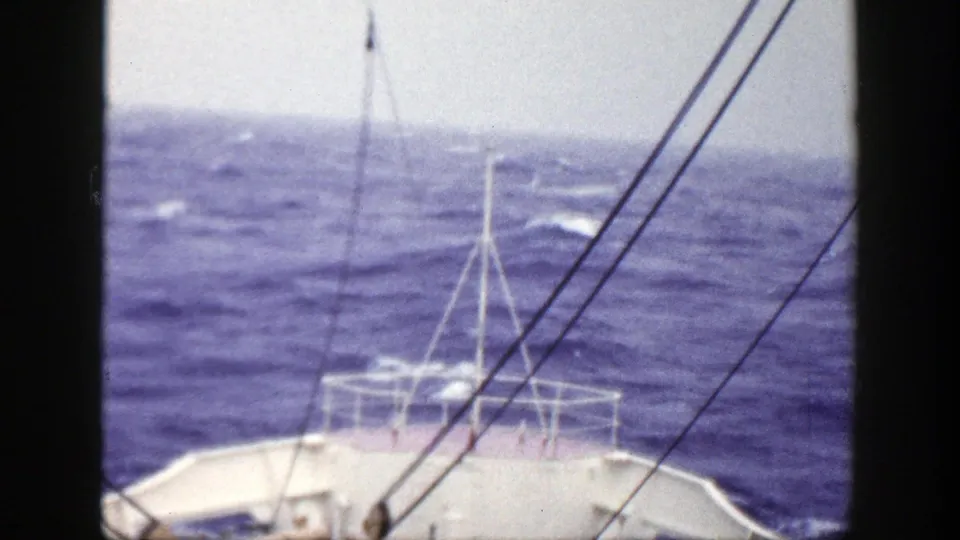 Vintage 8mm frame showing the boat's forward railing and diagonal rigging lines against blue water and a pale sky.