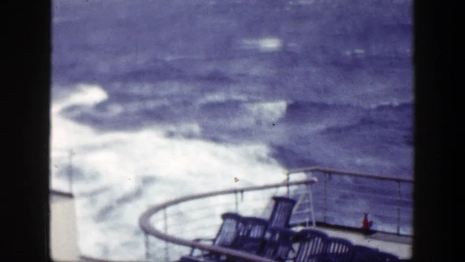 1948 8mm frame from a ship deck, with white wake, rough blue water, railing, and deck chairs visible.